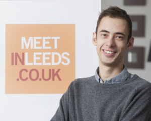 Corin is standing in front of a meet in leeds sign and is smiling.